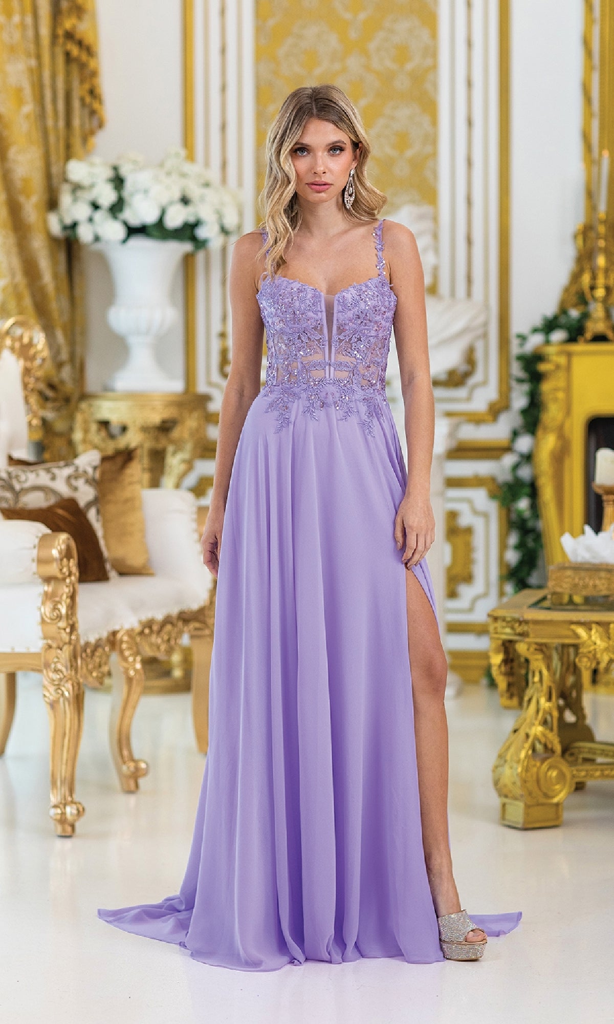 A woman wearing the Lace-Corset Long Prom Dress: Dancing Queen 4531 stands in an ornate room with gold and white decor and floral arrangements, showcasing the gown’s embroidered lace and front slit.